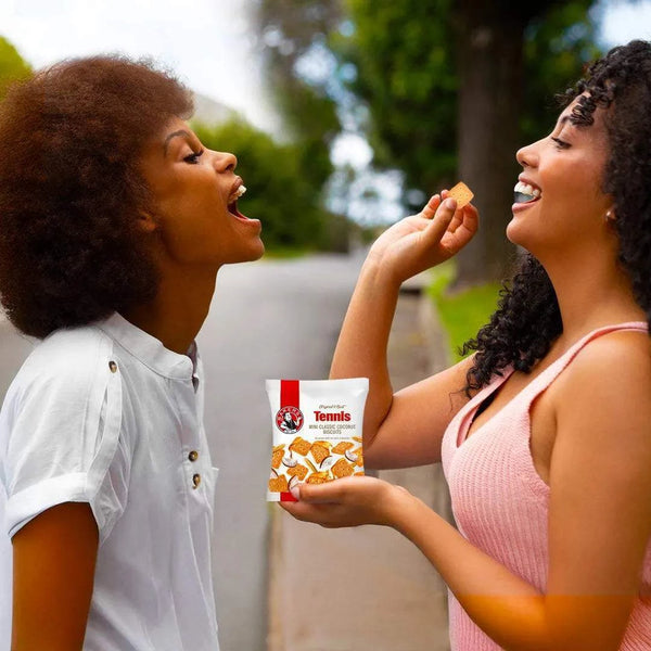 Two women outdoors sharing Bakers Tennis Mini Classic Coconut Biscuits 40g pack, enjoying a crunchy coconut snack