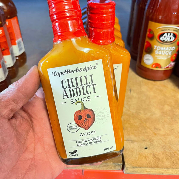 Hand holding a 200ml bottle of Cape Herb & Spice Chilli Addict Ghost Chilli Sauce with red cap and spicy ghost pepper label on wooden surface