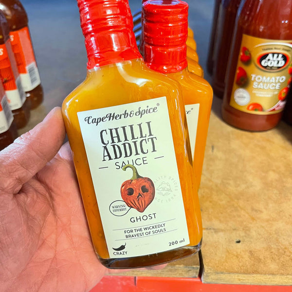Hand holding a 200ml bottle of Cape Herb & Spice Chilli Addict Ghost Chilli Sauce with red cap and spicy ghost pepper label on wooden surface