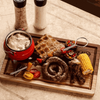 Traditional 3-legged cast iron pot with rustic meal, featuring sausage, corn, and grilled bread on a wooden board.