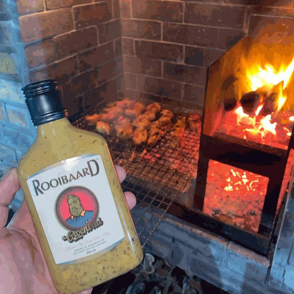 Rooibaard Groen Trui Chilli Sauce 250ml bottle held near a barbecue grill with meat cooking.