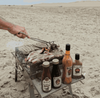 Outdoor barbecue with Rooibaard Sauce bottles on a sandy beach.