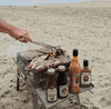 Outdoor barbecue with Rooibaard Sauce bottles on a sandy beach.