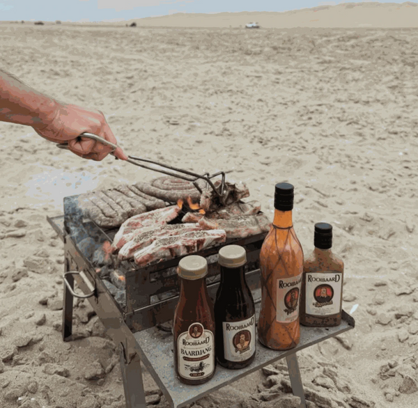 Outdoor barbecue with Rooibaard Sauce bottles on a sandy beach.