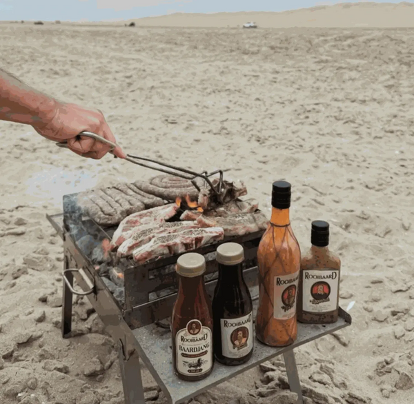 Outdoor barbecue with Rooibaard Sauce bottles on a sandy beach.