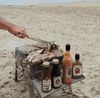 Outdoor barbecue with Rooibaard Sauce bottles on a sandy beach.