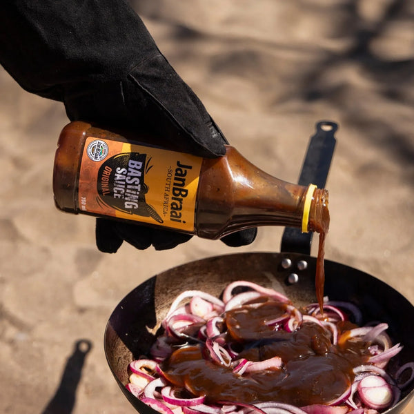 Jan Braai Basting Original sauce poured over sliced onions in a pan, South African BBQ flavor, 750ml bottle