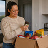 Grocery delivery box with South African Shops NZ pantry products, including Milo and KOO, held by a woman in a cream knit sweater.