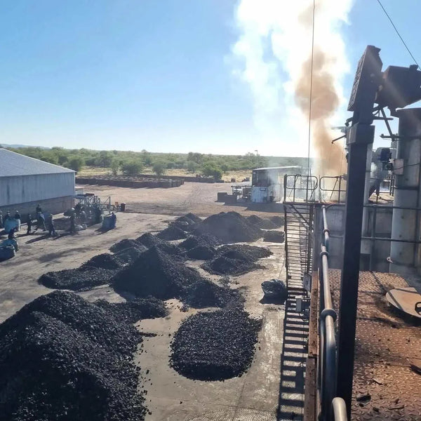 Industrial charcoal production site with large piles of black charcoal and machinery emitting smoke under a clear sky