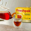 Freshpak Rooibos Tea pouring into a glass cup with vibrant red tea and Bright Freshpak Rooibos Tea 80's box in background