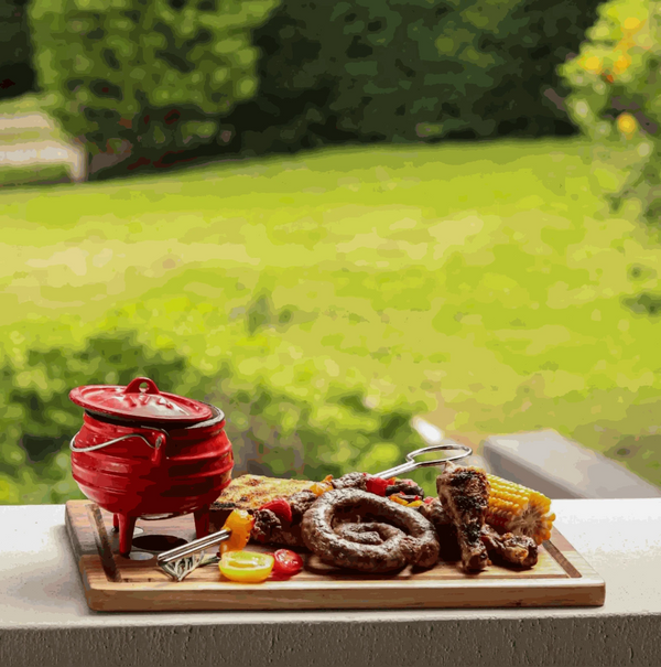 Traditional 3-legged red enamel cast iron pot with lid, surrounded by barbecue food, perfect for outdoor cooking.