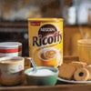 Ricoffy 750g tin with coffee and biscuits on a table.