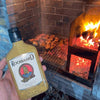 Rooibaard Groen Trui Chilli Sauce 250ml bottle held near a barbecue grill with meat cooking.